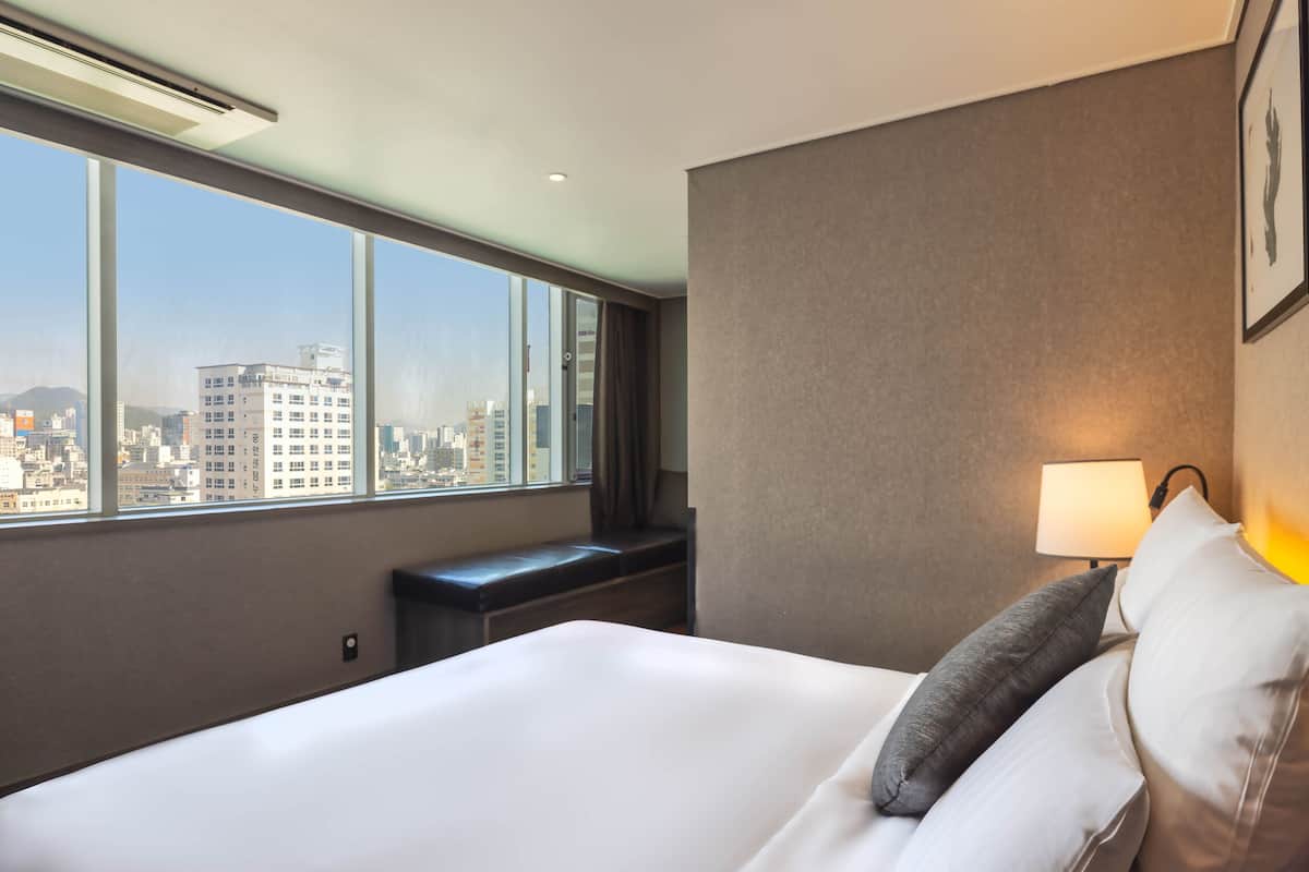 Une chambre d’hôtel avec un grand lit, une banquette, une fenêtre donnant sur la ville et une lampe fixée au mur.