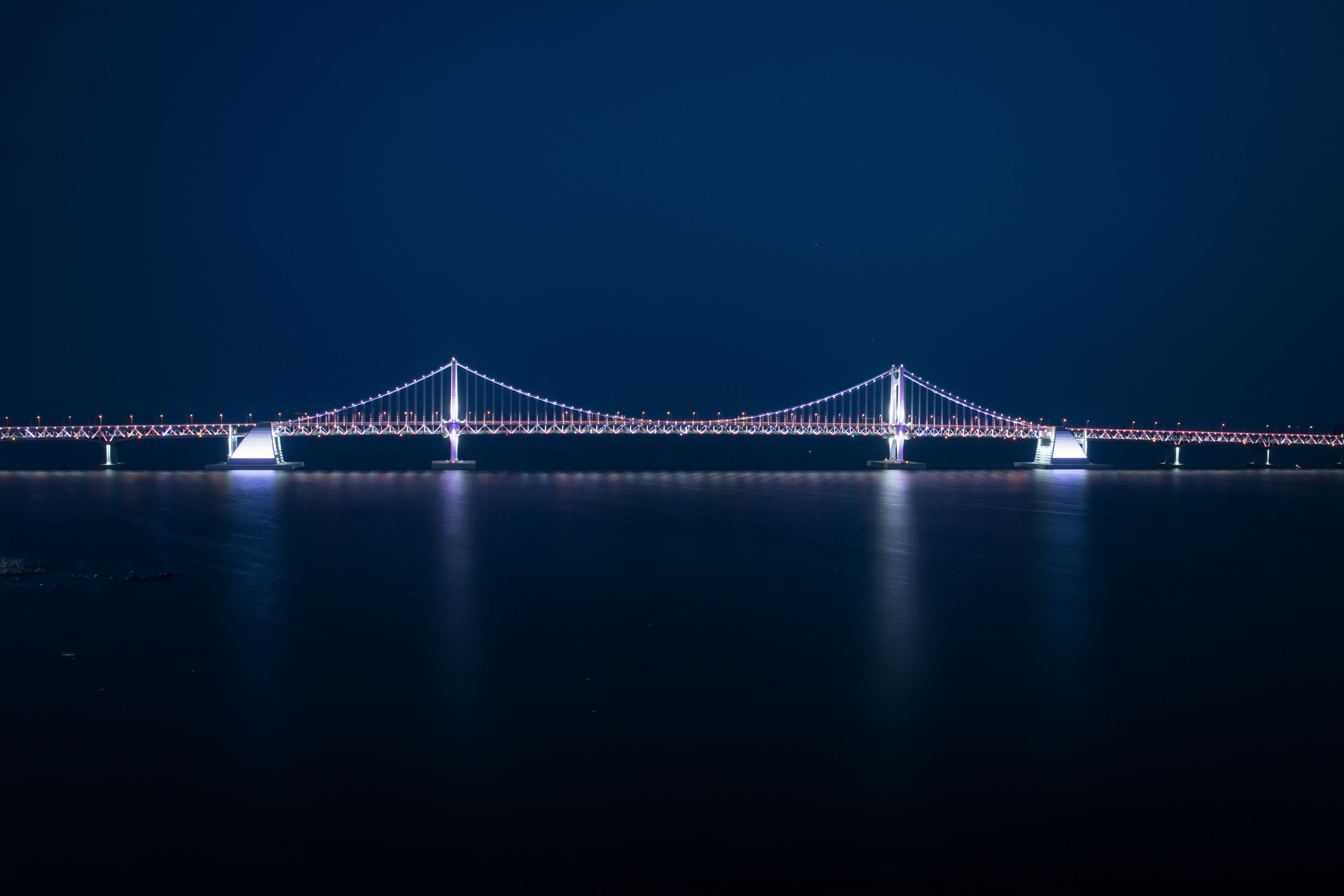 A nighttime view of a large suspension bridge with illuminated cables and towers, spanning over a body of water.