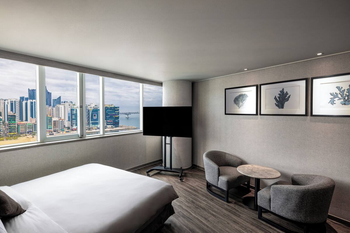 Une chambre d’hôtel avec un lit, deux fauteuils, une petite table, un téléviseur et des œuvres d’art encadrées accrochées au mur.