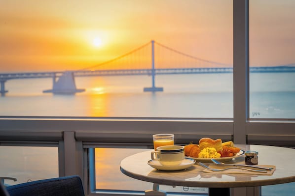 Una mesa de desayuno con una taza, vaso, tostada y mermelada, con vistas a un puente al atardecer.