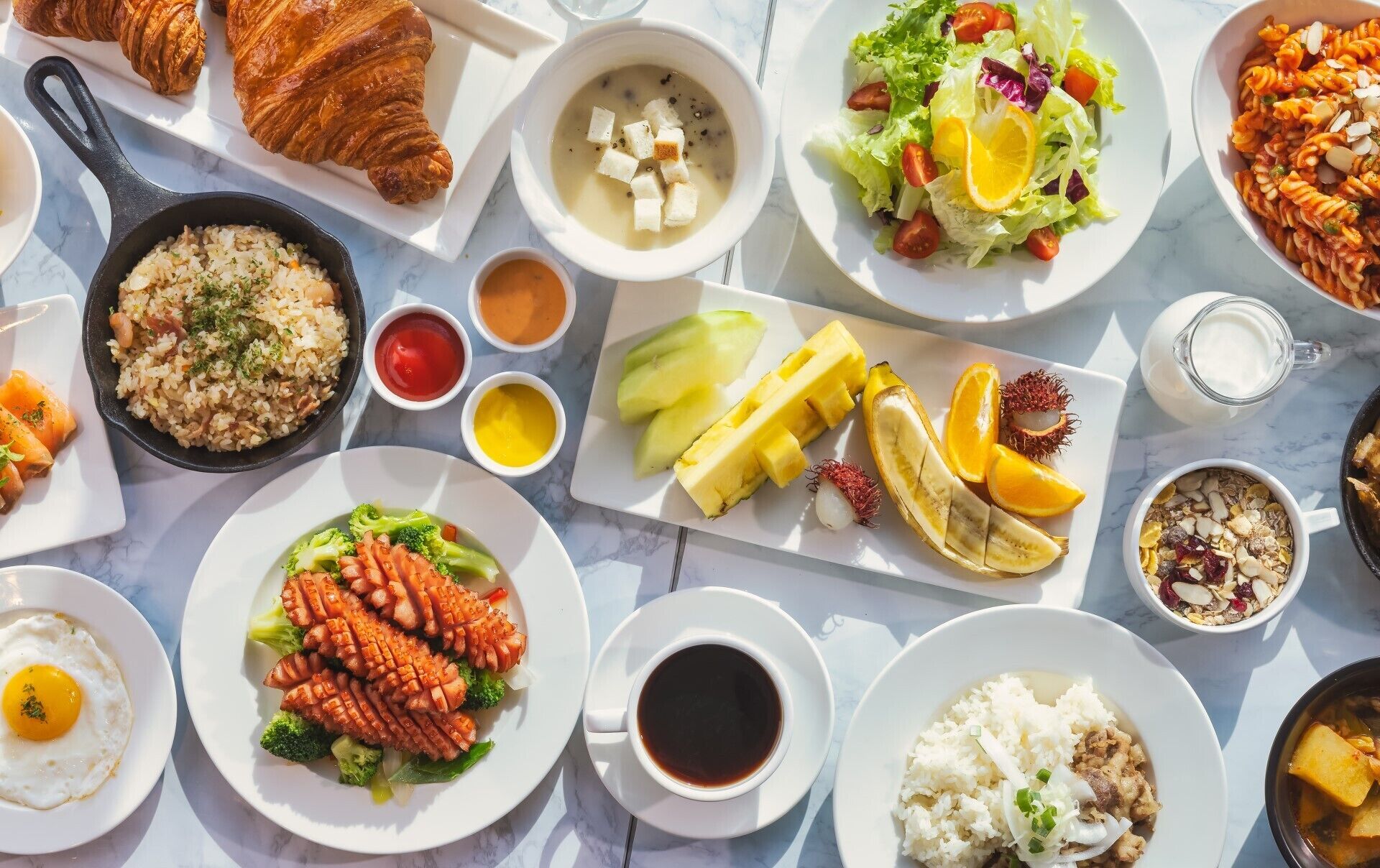 A variety of dishes including croissants, rice, and fruit on a marble table.