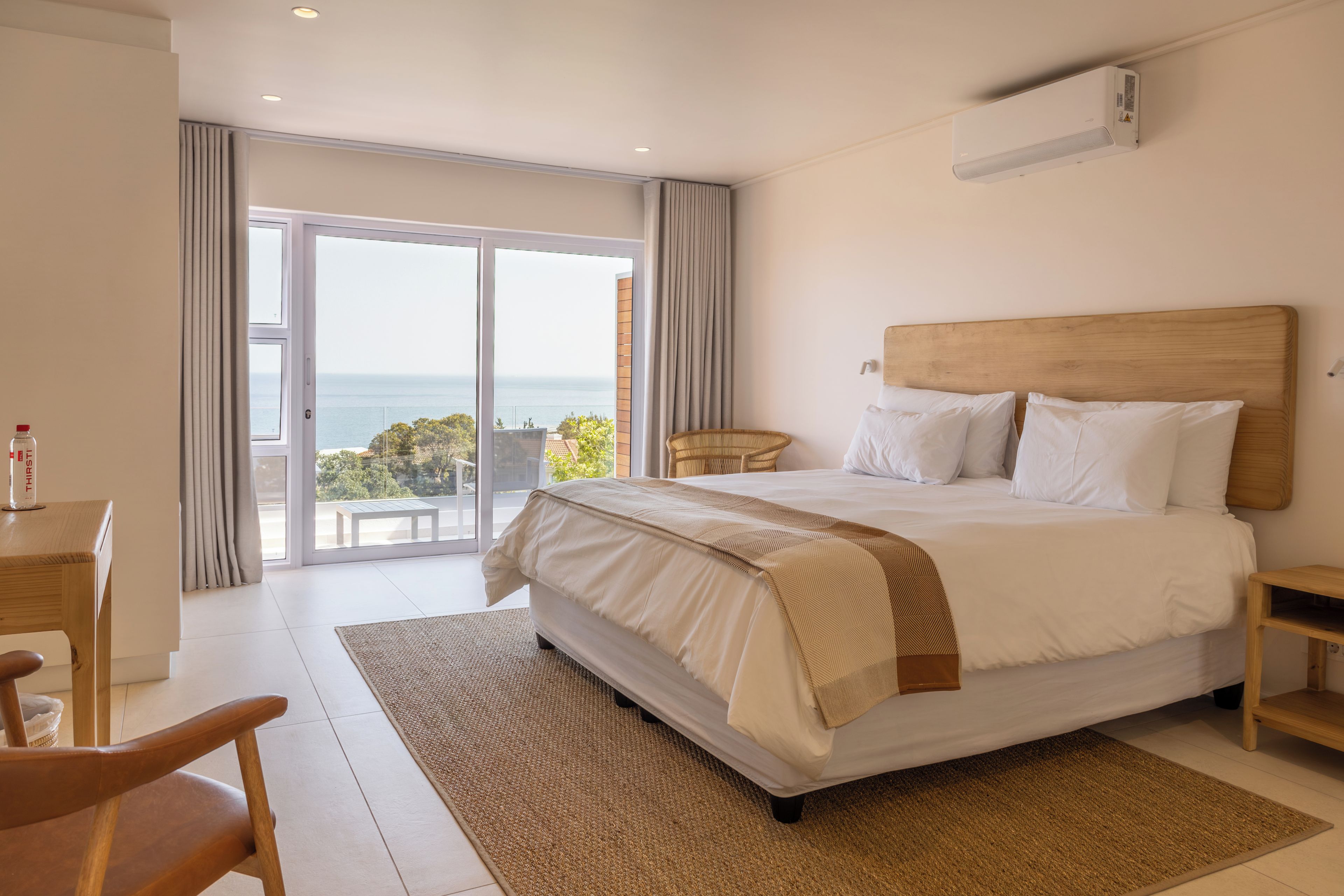 Ein Hotelzimmer mit einem großen Bett, einem Holz-Kopfteil, einem kleinen Nachttisch, einem Sessel und Meerblick durch eine Glastür.