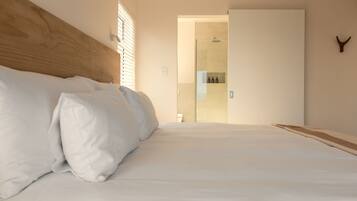 Quarto de hotel com uma cama grande, cabeceira de madeira, banheiro com porta de vidro e um cabideiro.