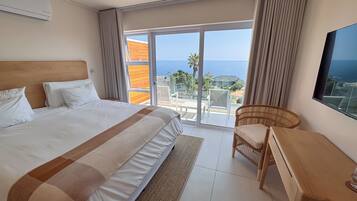 A hotel room with a large bed, a desk, a chair, and a view of the sea through a sliding glass door.