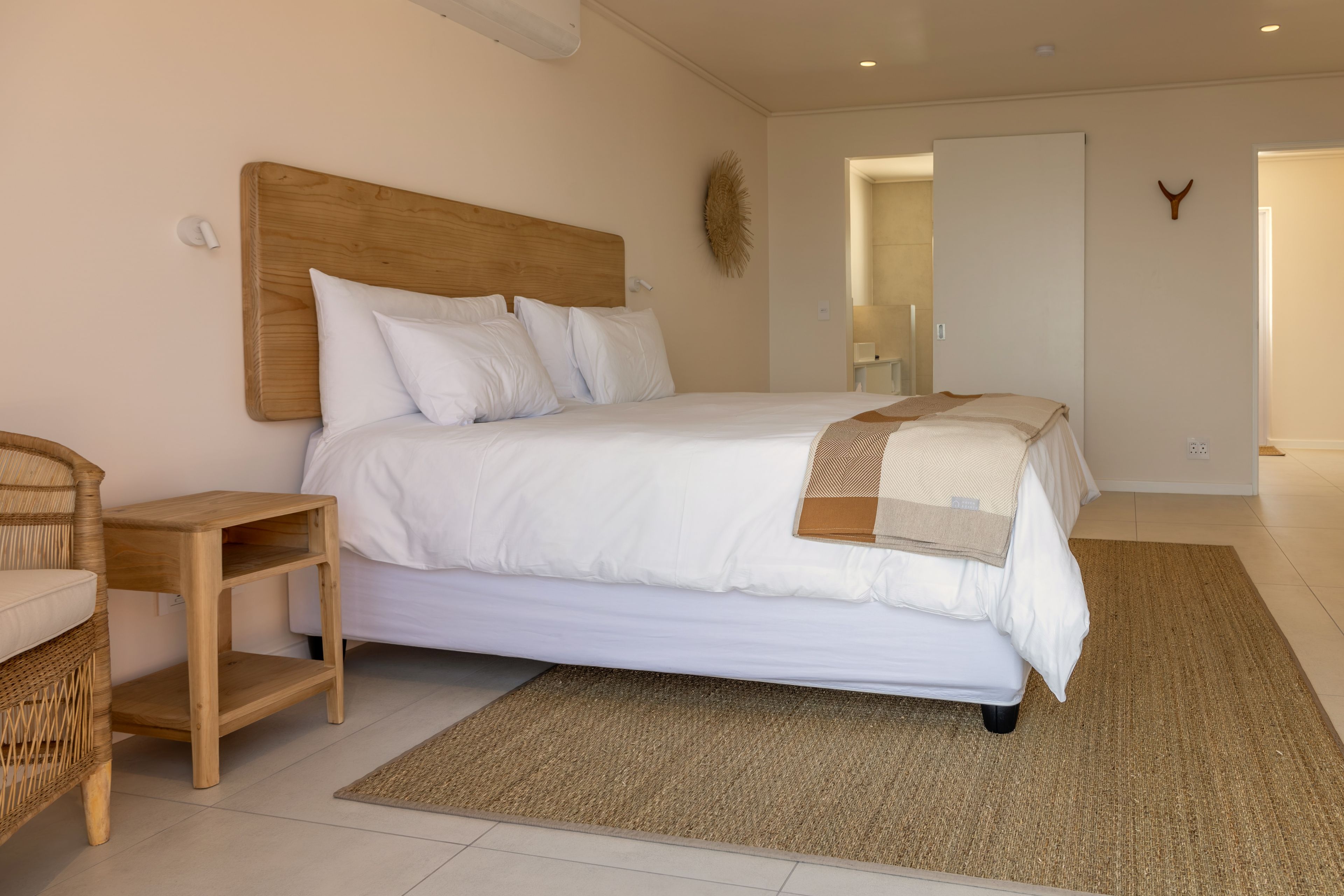 Una camera da letto moderna con una testiera in legno, biancheria da letto bianca, un tappeto tessuto e una sedia in vimini.