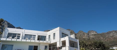 A modern white building with multiple balconies and large windows, labeled 'Finchley Guesthouse,' set against a mountainous backdrop.