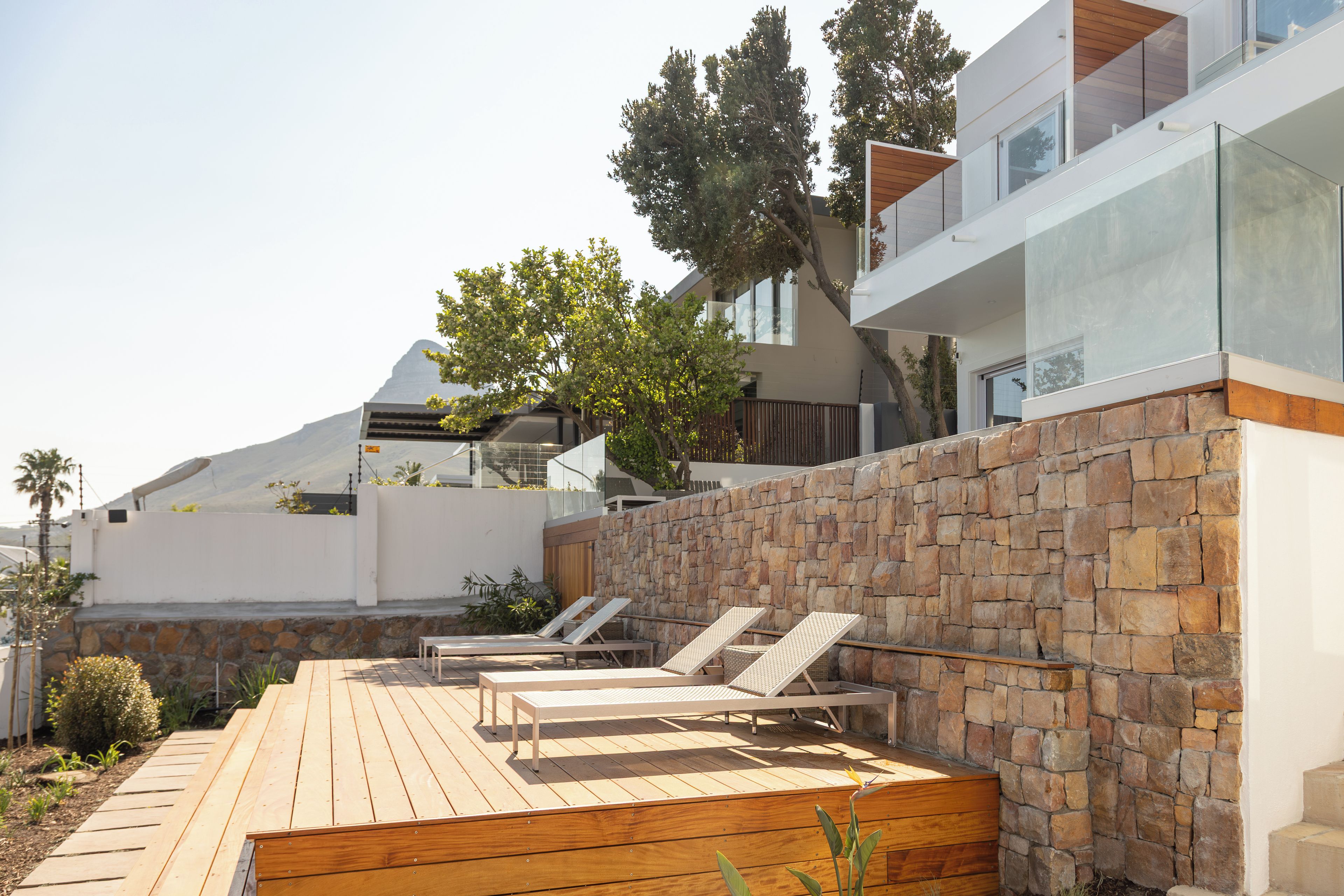 A modern outdoor area with a wooden deck, lounge chairs, and a stone wall.