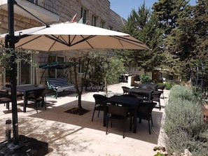 An outdoor patio area with tables and chairs, a large umbrella, and a stone building in the background. - Talitha Kumi Guest House (Bethlehem)