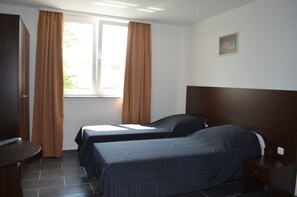 Una habitación de hotel con dos camas individuales, cabecero de madera, una ventana con cortinas, una mesita y un cuadro colgado en la pared.