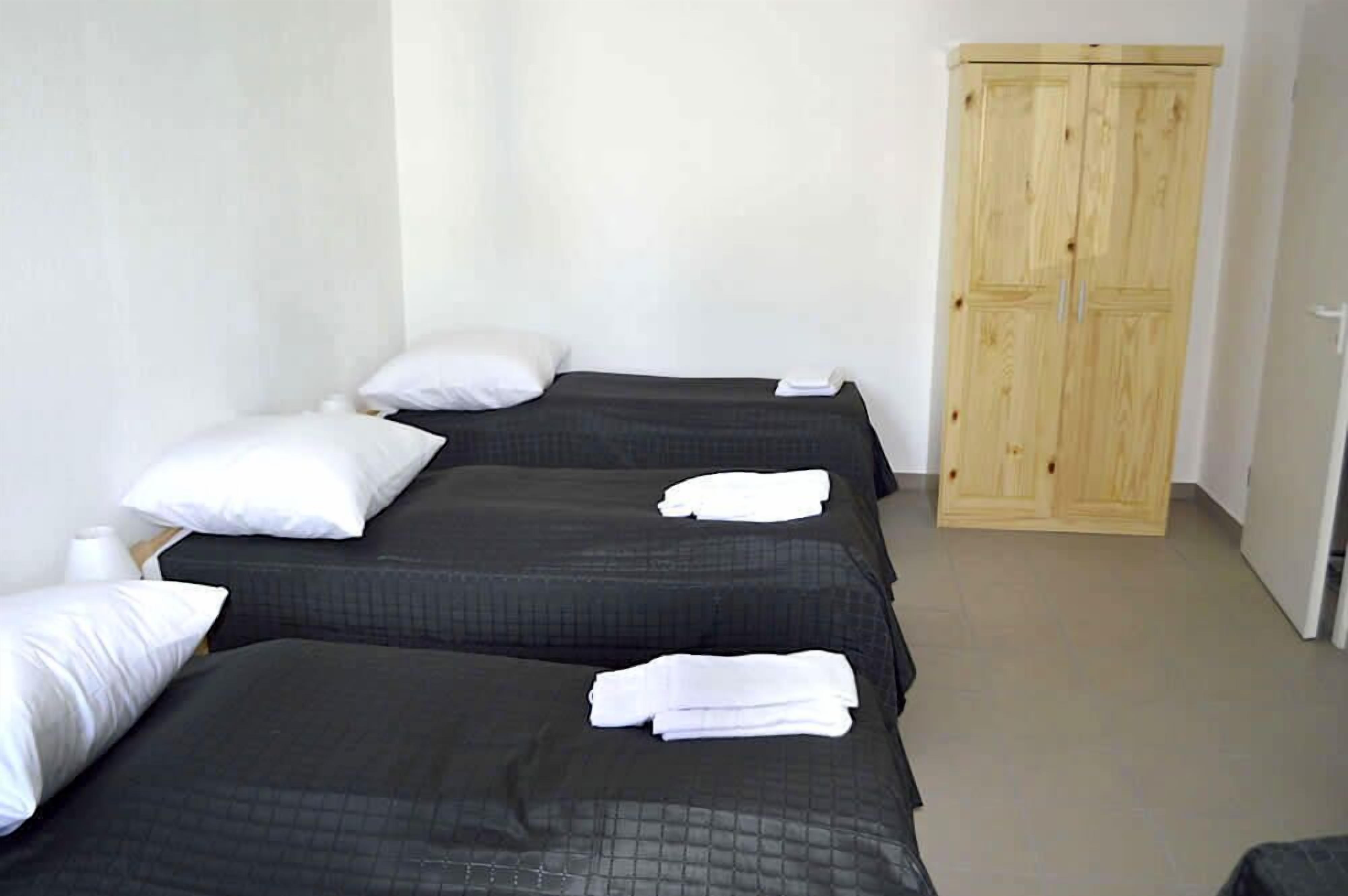 Una habitación con dos camas, cada una con un edredón oscuro y almohadas blancas. Se observa un armario de madera adosado a la pared.