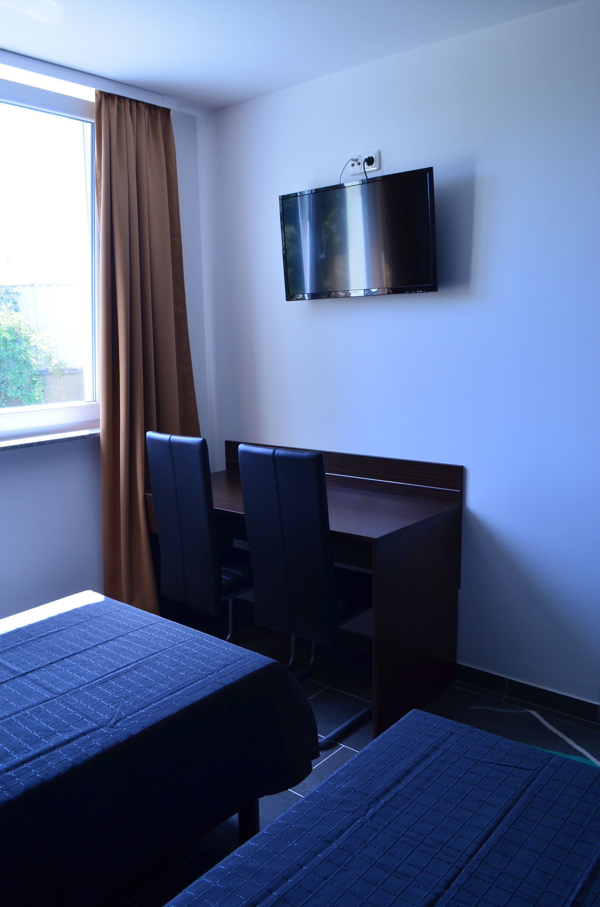 Habitación de hotel con cama, escritorio, dos sillas, televisor montado en la pared y ventana con cortinas.