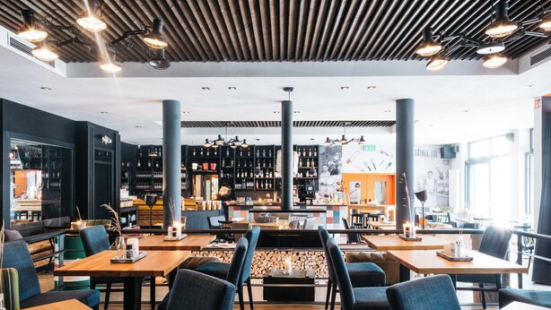 A modern restaurant with wooden tables, blue chairs, and a bar area with shelves of bottles. - Krögers Hotel (Esens)