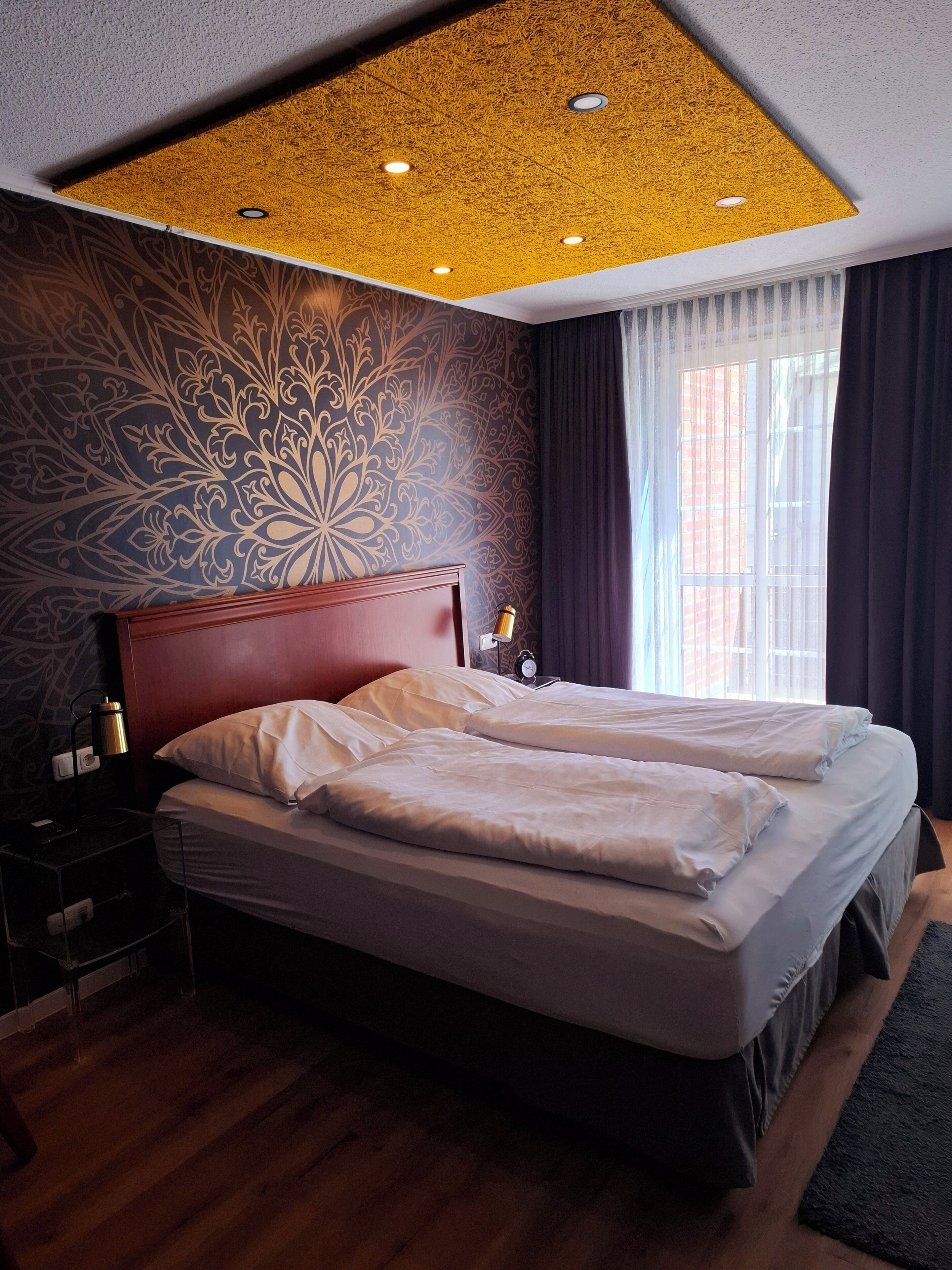 A hotel room with a large bed, a wooden headboard, a patterned wall, and a ceiling with recessed lighting.
