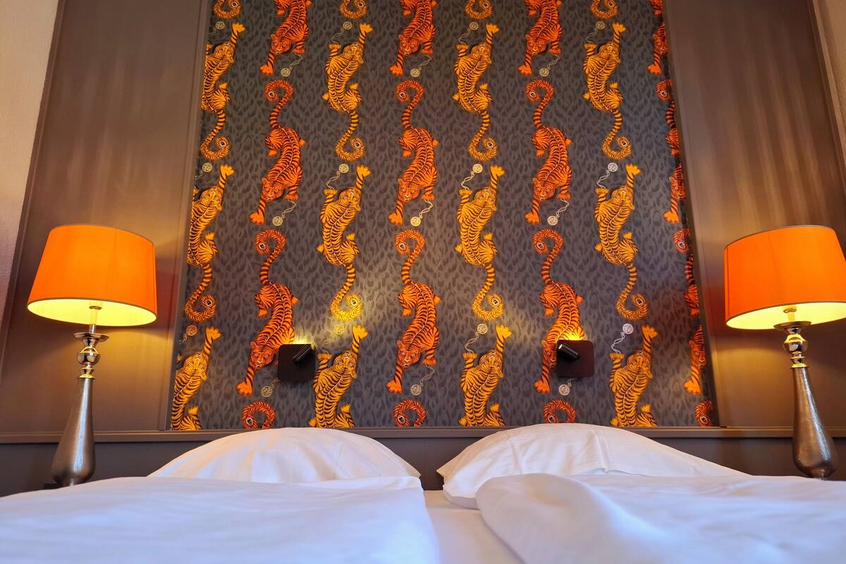 A hotel room with a patterned wallpaper, two bedside lamps, and a bed with white linens.