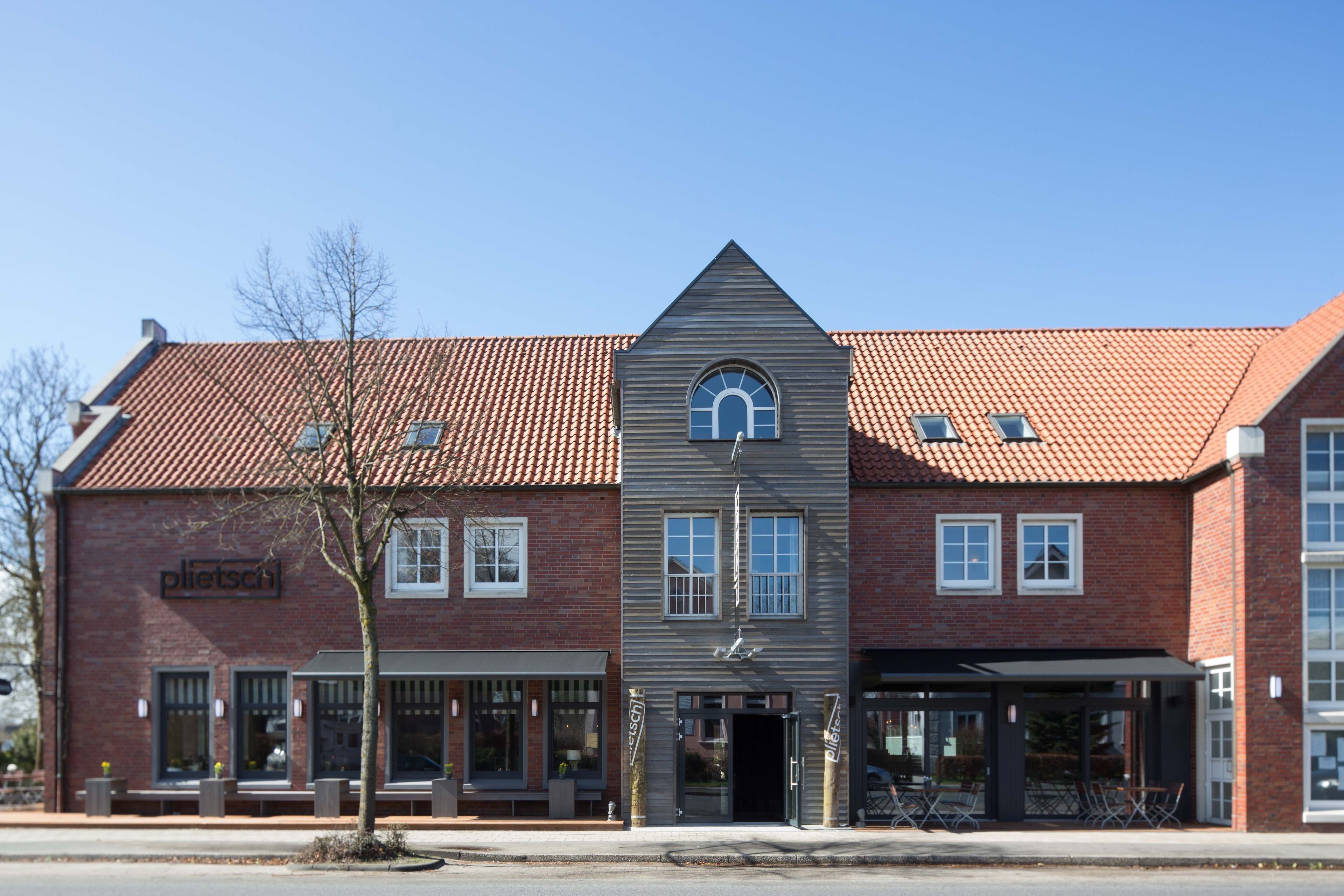 A two-story building with a red-tiled roof, a central entrance with a triangular pediment, and a sign reading "plietscht."