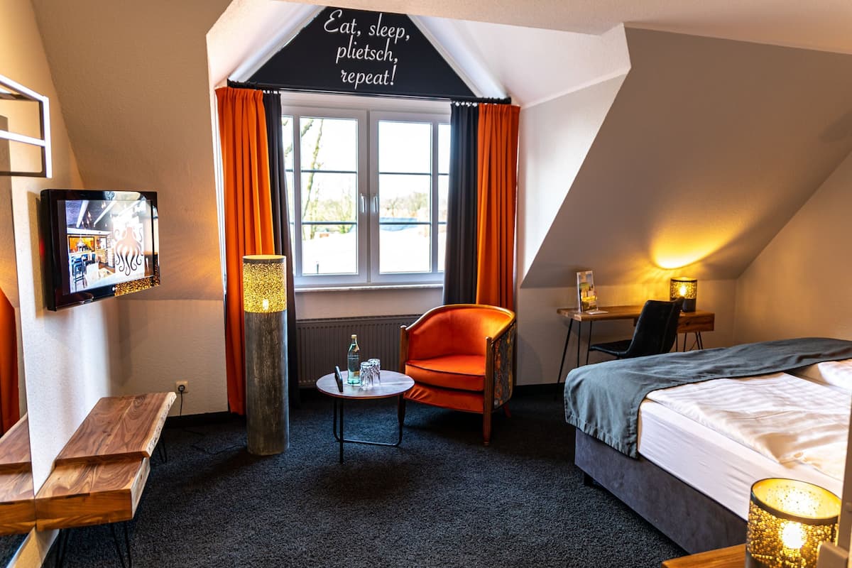A hotel room with a bed, a chair, a small table, a TV, and a window with curtains.