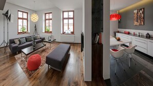 A modern living room with a sofa, coffee table, and ottoman, a kitchen with white cabinets and red pendant lights, and large windows with plants. - Apartament ST1 (Cieszyn)