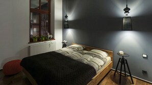 A modern bedroom with a wooden bed, a window with plants, a red pouf, and a small round table with a cup. - Apartament ST1 (Cieszyn)