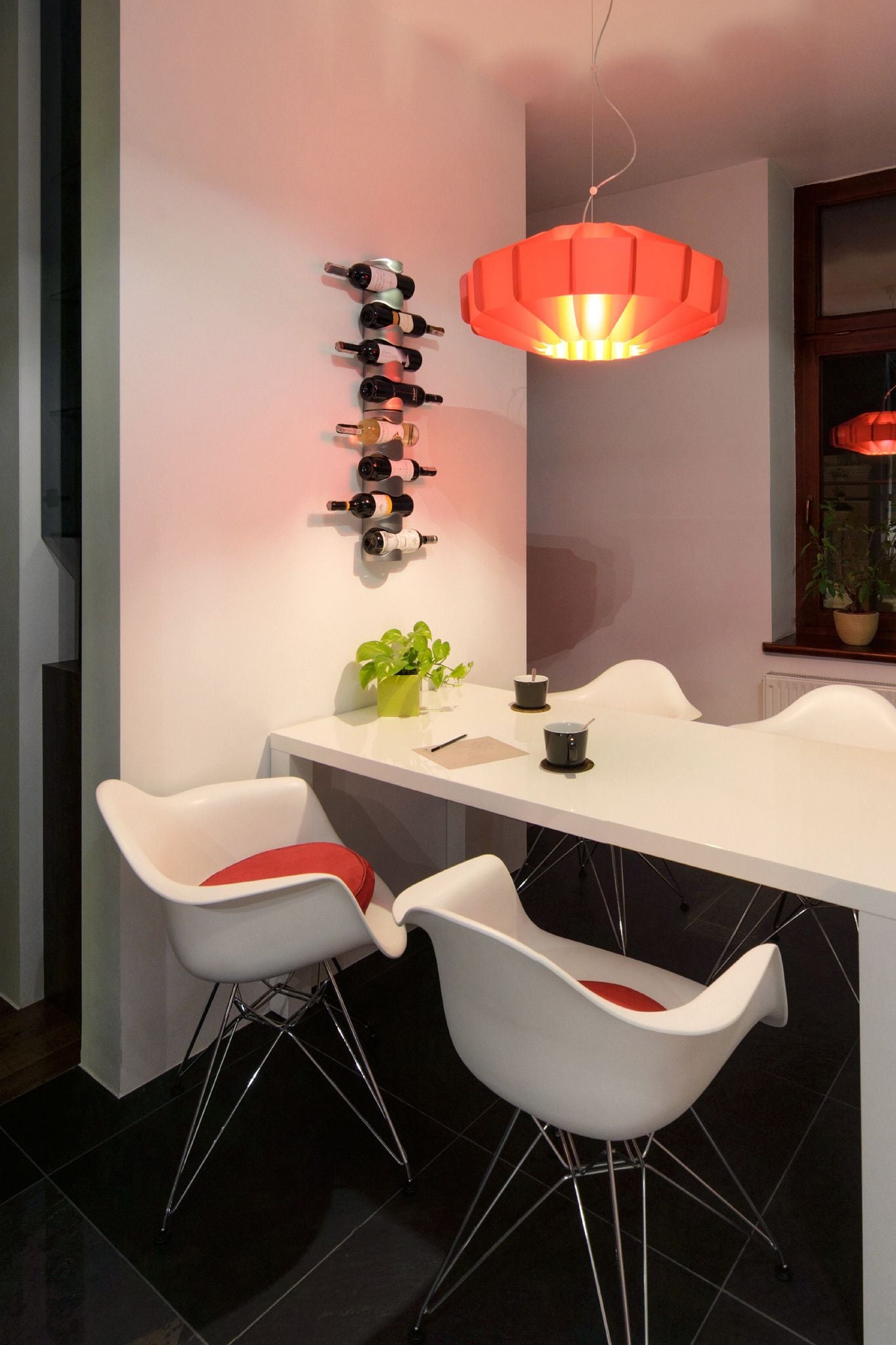 A modern dining area with a white table, two white chairs with red cushions, a pendant light, a wall-mounted wine rack, and a potted plant.