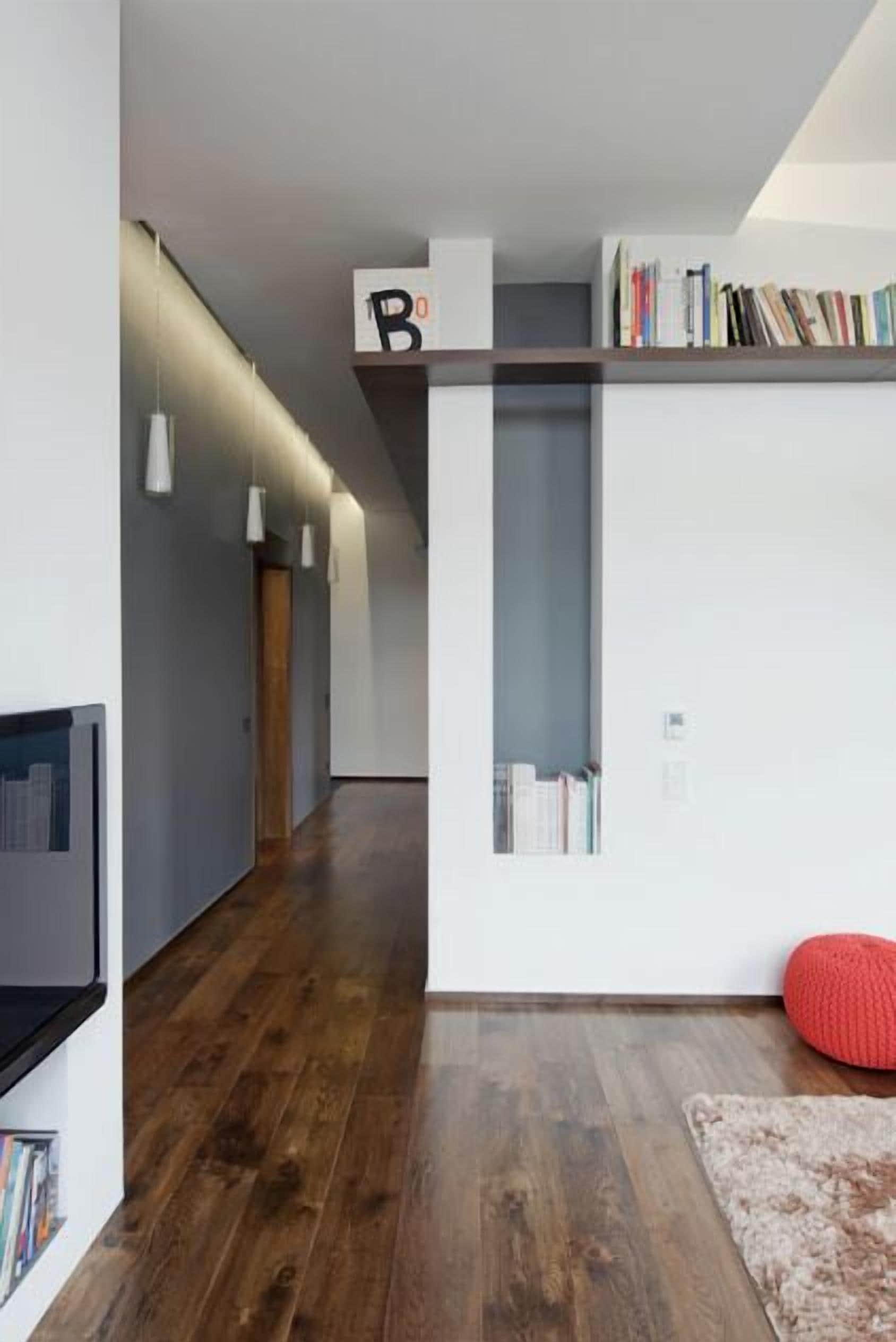 Ein modernes Interieur mit Holzboden, einer Bücherwand, einem roten Pouf und einem an der Wand befestigten Bücherregal.