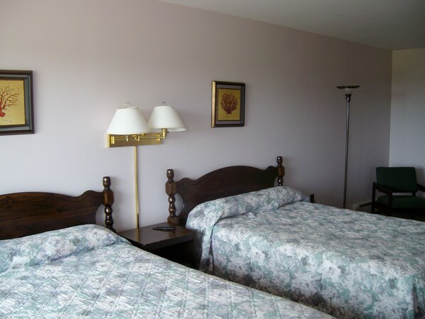 A hotel room with two single beds, a nightstand with a lamp, a chair, and framed artwork on the wall. - Singing Sands Inn Motel (Kingsboro)