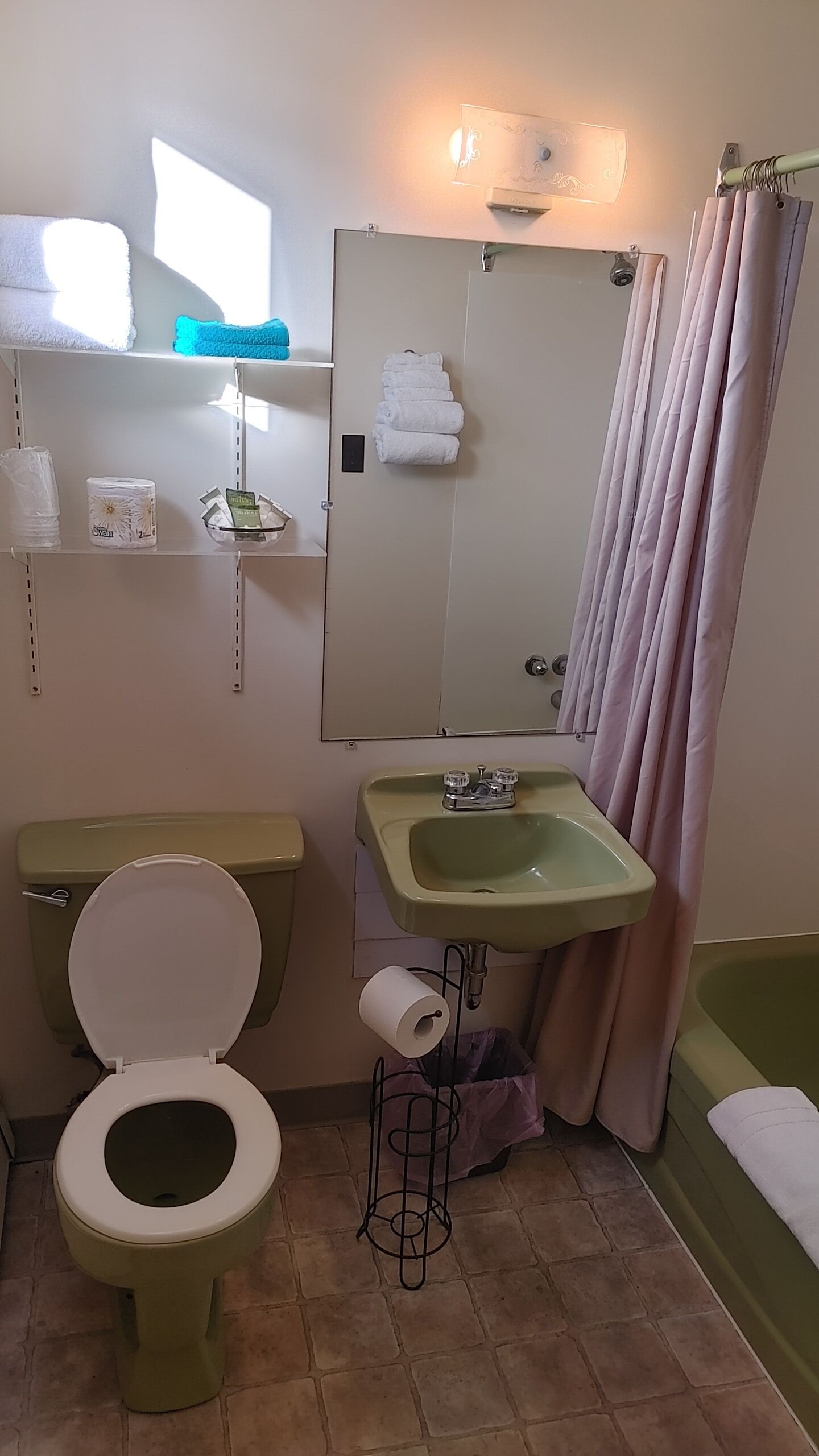 Baño con inodoro, lavamanos y bañera. Hay toallas y artículos de aseo en estantes.