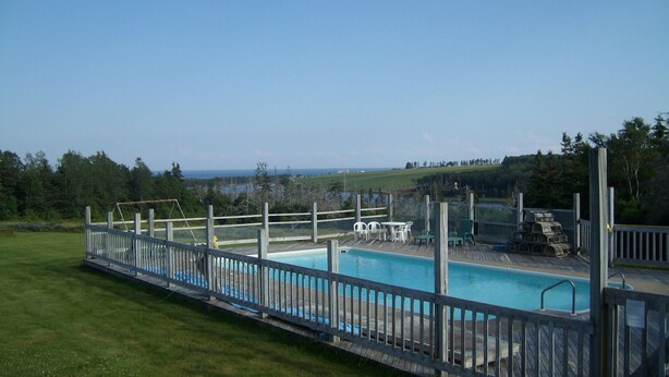 A rectangular swimming pool with a wooden deck and a fence, surrounded by grass and trees. - Singing Sands Inn Motel (Kingsboro)