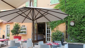 Une terrasse extérieure avec des tables et des chaises, un grand parasol et un bâtiment aux volets verts et recouvert de lierre.