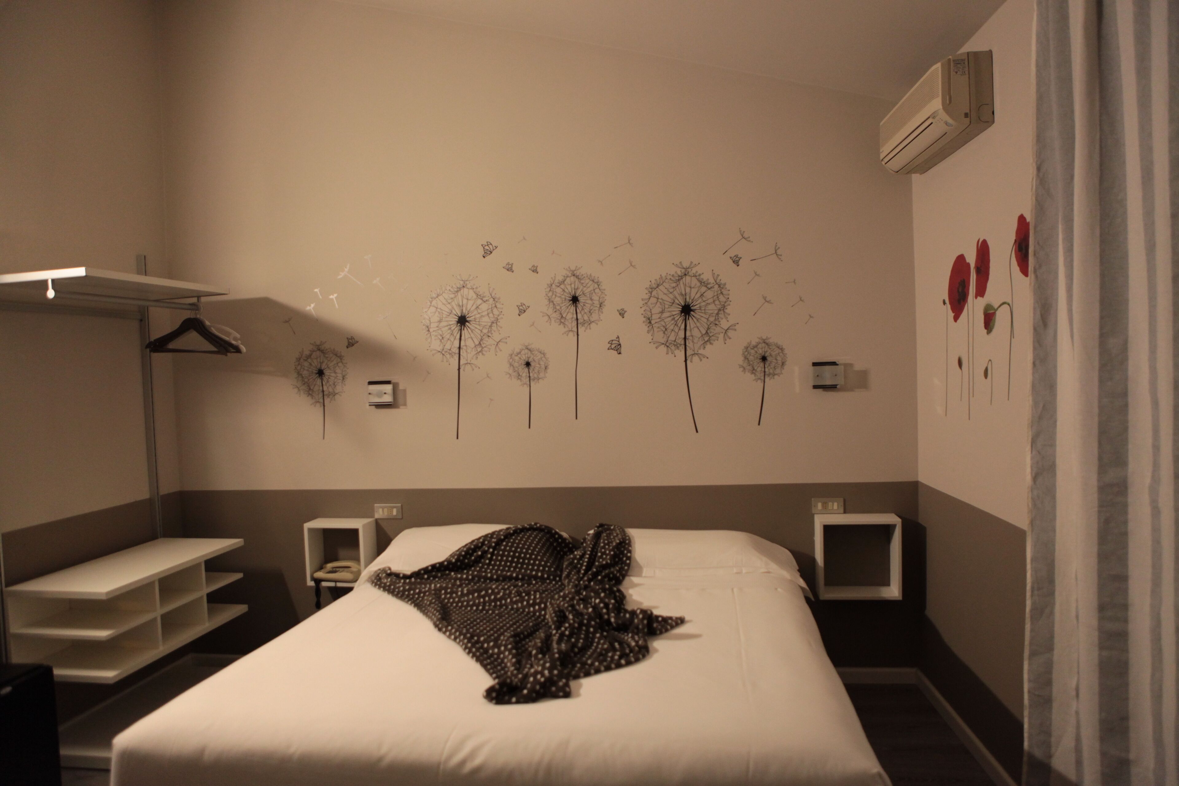 A hotel room with a bed, a wardrobe, a bedside table, a wall-mounted air conditioner, and a decorative wall with dandelion and poppy motifs.