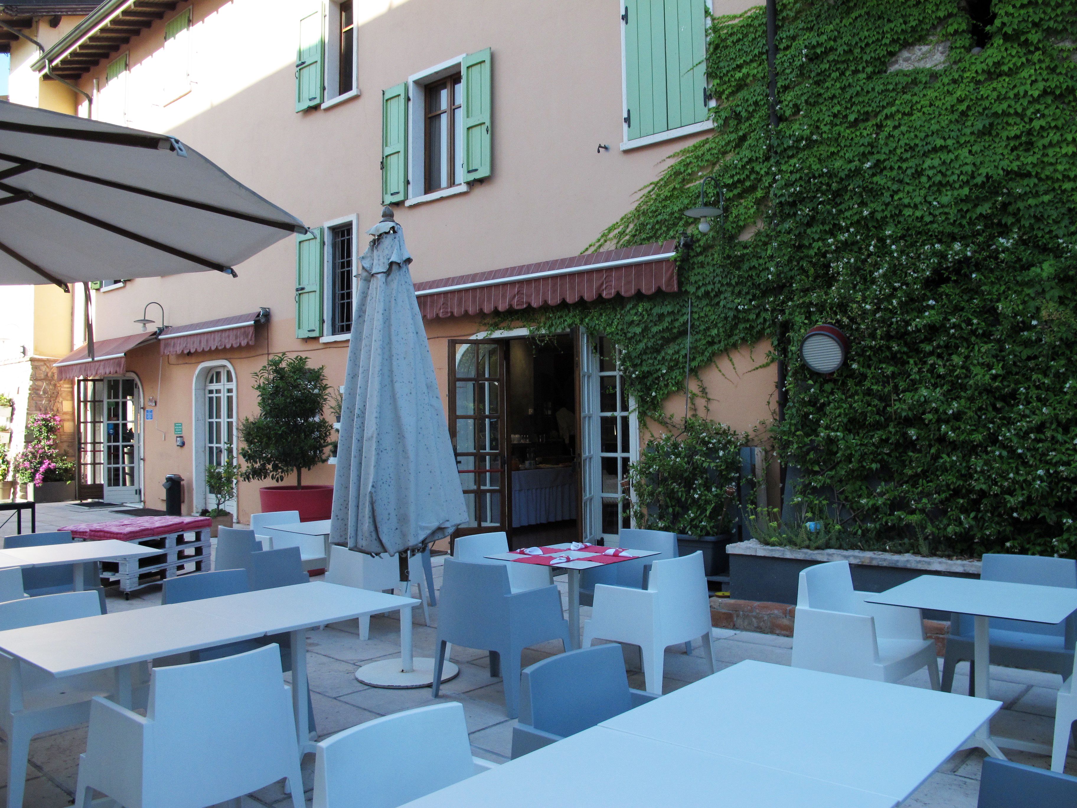 An outdoor café with white tables and chairs, a covered umbrella, and a building with green shutters and ivy.