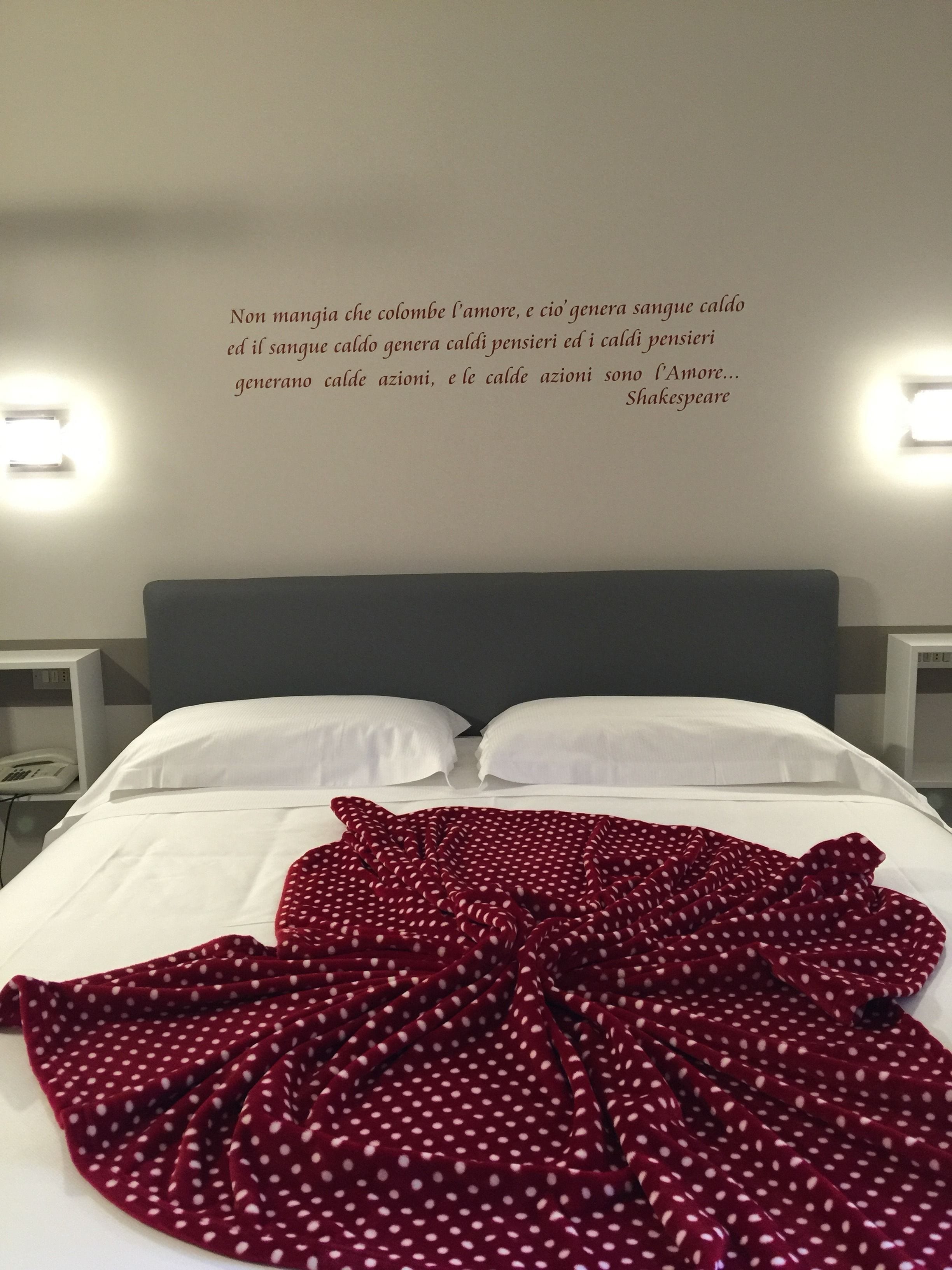 Un letto rifatto con lenzuola bianche e una coperta rossa a pois. Ci sono due comodini con lampade e una citazione sulla parete sopra il letto.