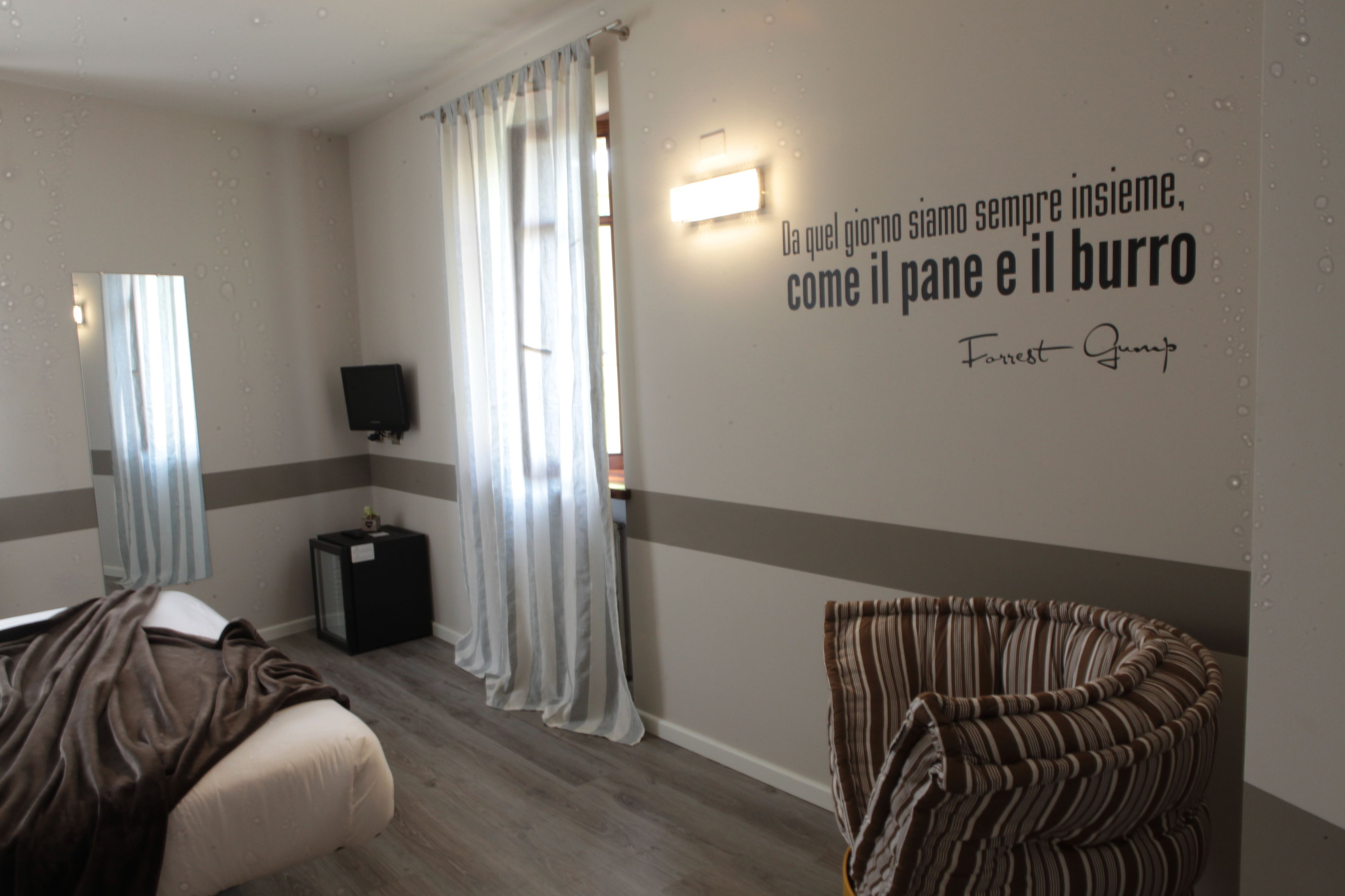 A hotel room with a bed, a TV, a wall-mounted light, a window with curtains, and a quote by Forrest Gump on the wall.