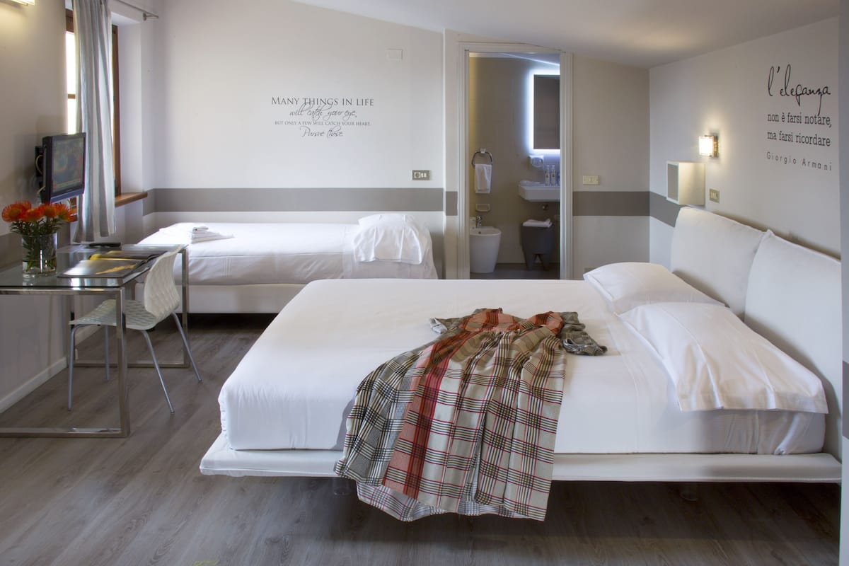 A hotel room with two beds, a desk with a chair, a small table with a vase of flowers, and a bathroom visible through an open door.