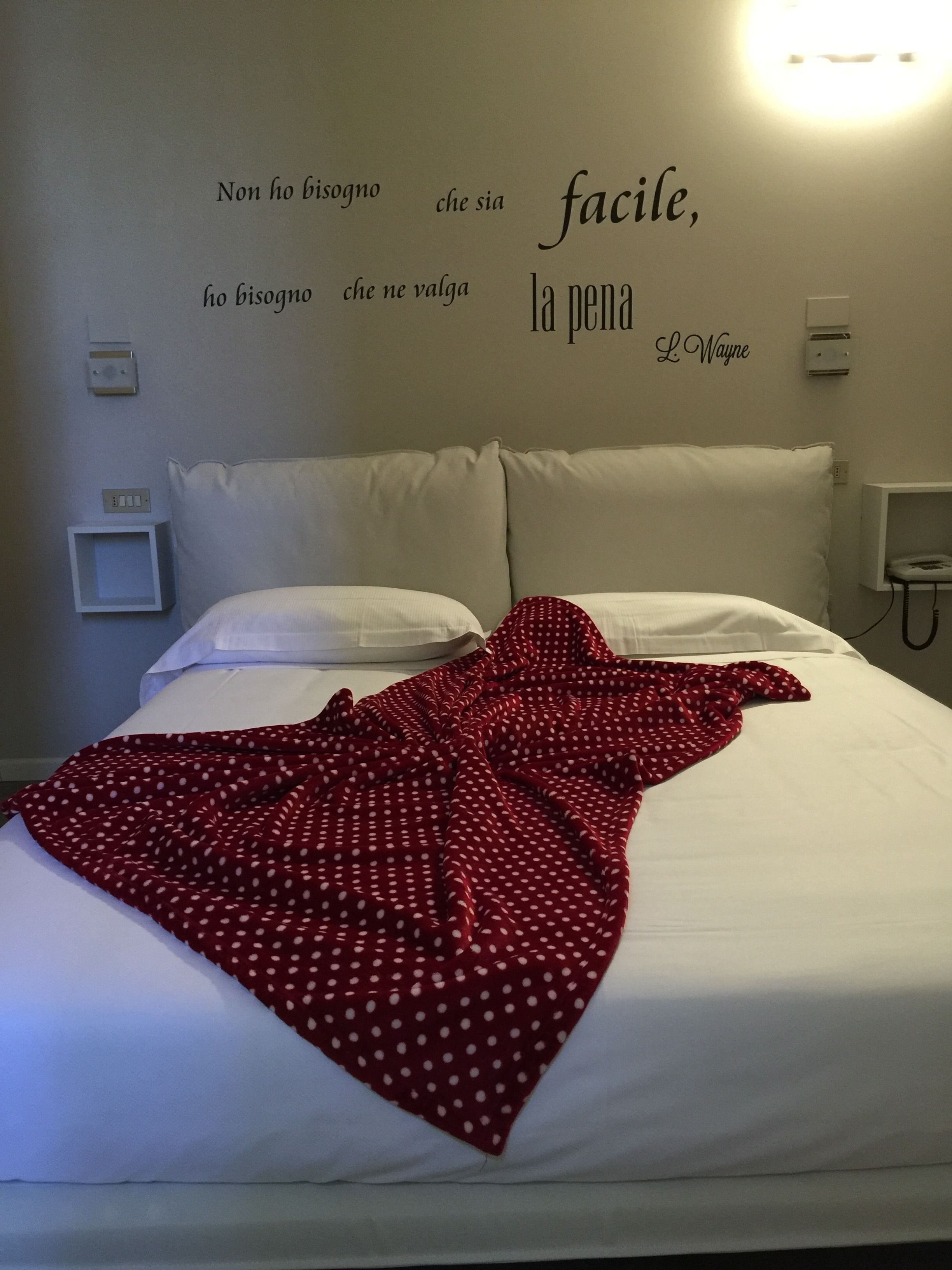 Ein ordentlich bezogenes Bett mit einer puntierten Decke, weißen Kissen und einer Wand mit einem motivierenden Zitat auf Italienisch.