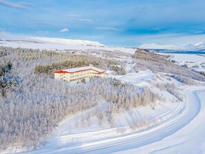 A snow-covered landscape with a building featuring a red roof and multiple windows, surrounded by trees and a winding path.
