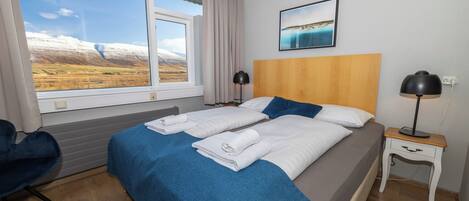 Ein Hotelzimmer mit großem Fenster, einem Bett mit blauer Bettwäsche, einem hölzernen Kopfteil, einem Nachttisch mit Lampe und einem Sessel.