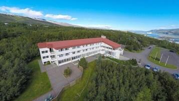 A white building with a red roof, surrounded by dense forest, with a parking area and a view of distant mountains.