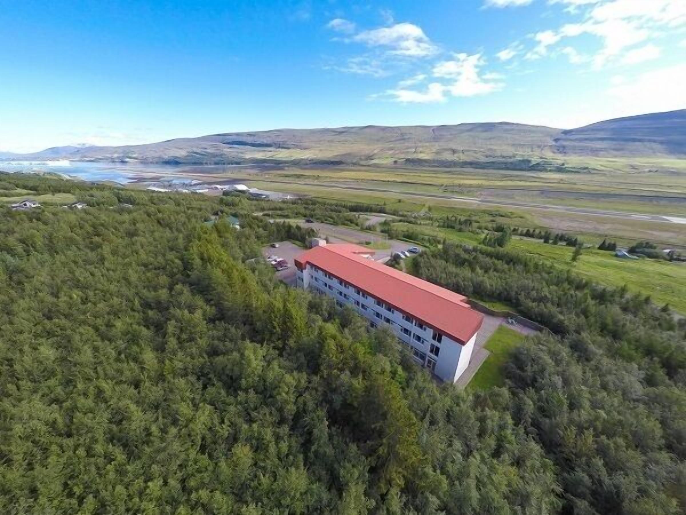 An aerial view of a building with a red roof, surrounded by dense forest and a river in the background.