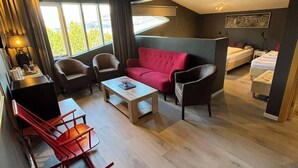 A modern, compact living space with a red sofa, a wooden coffee table, and a red rocking chair. There is a bed with a white mattress and a small bedside table. The room has large windows allowing natural light and a view of greenery outside. - Hotel Kjarnalundur (Akureyri)