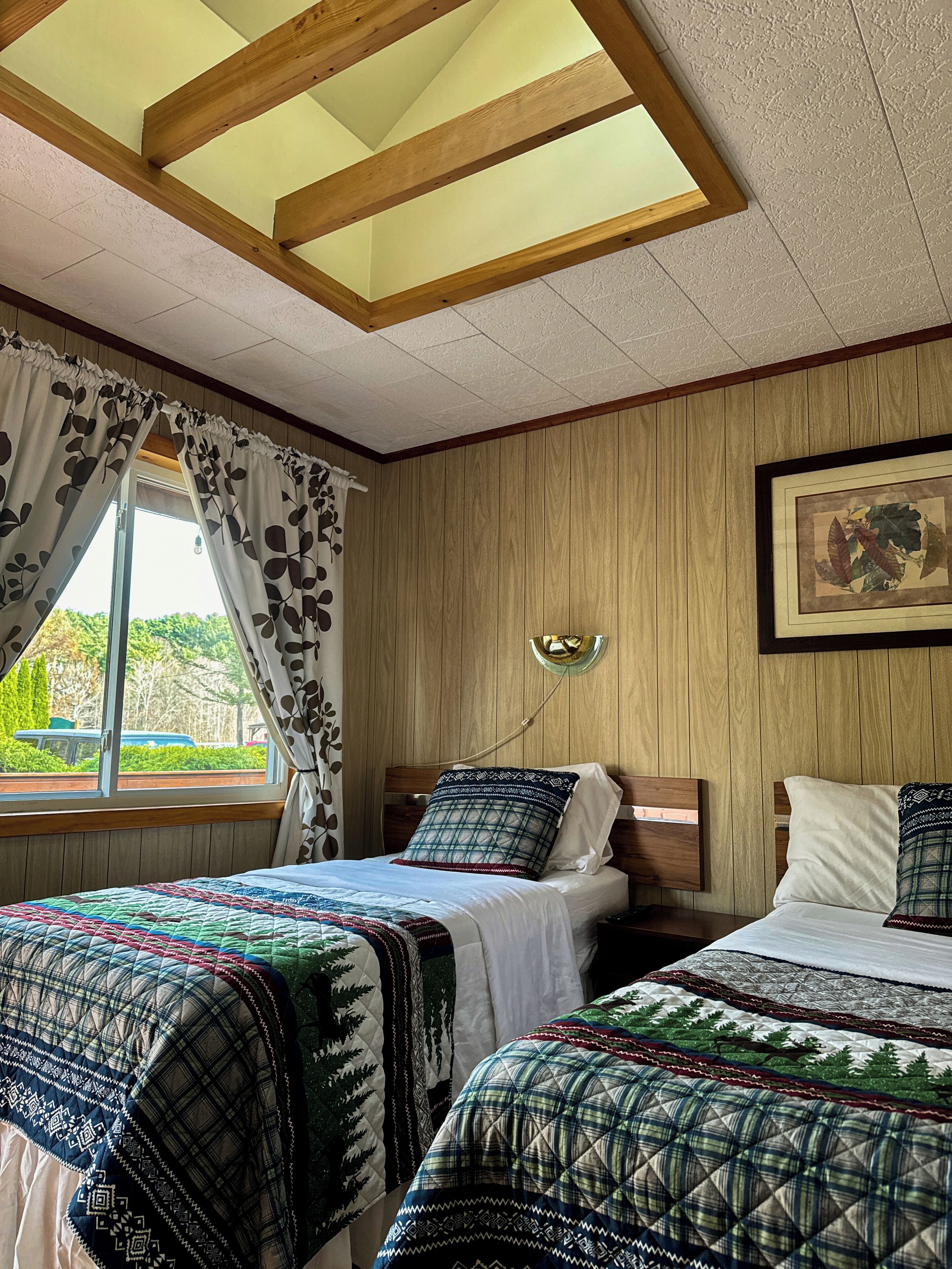 A bedroom with two beds, wooden panel walls, a skylight, and a window with curtains.
