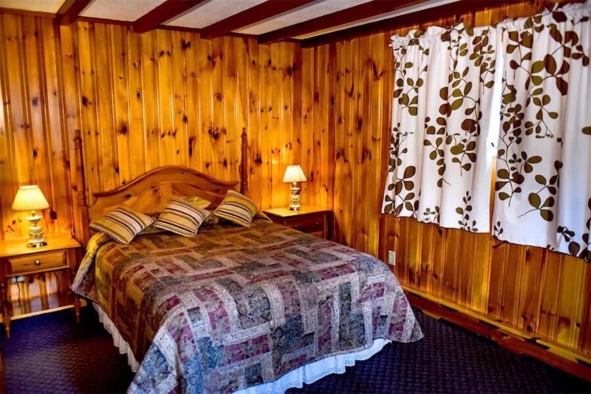 Ein Schlafzimmer mit Holzwandverkleidung, einem Bett, zwei Nachttischen mit Lampen und einem Fenster mit blattgemusterten Vorhängen.