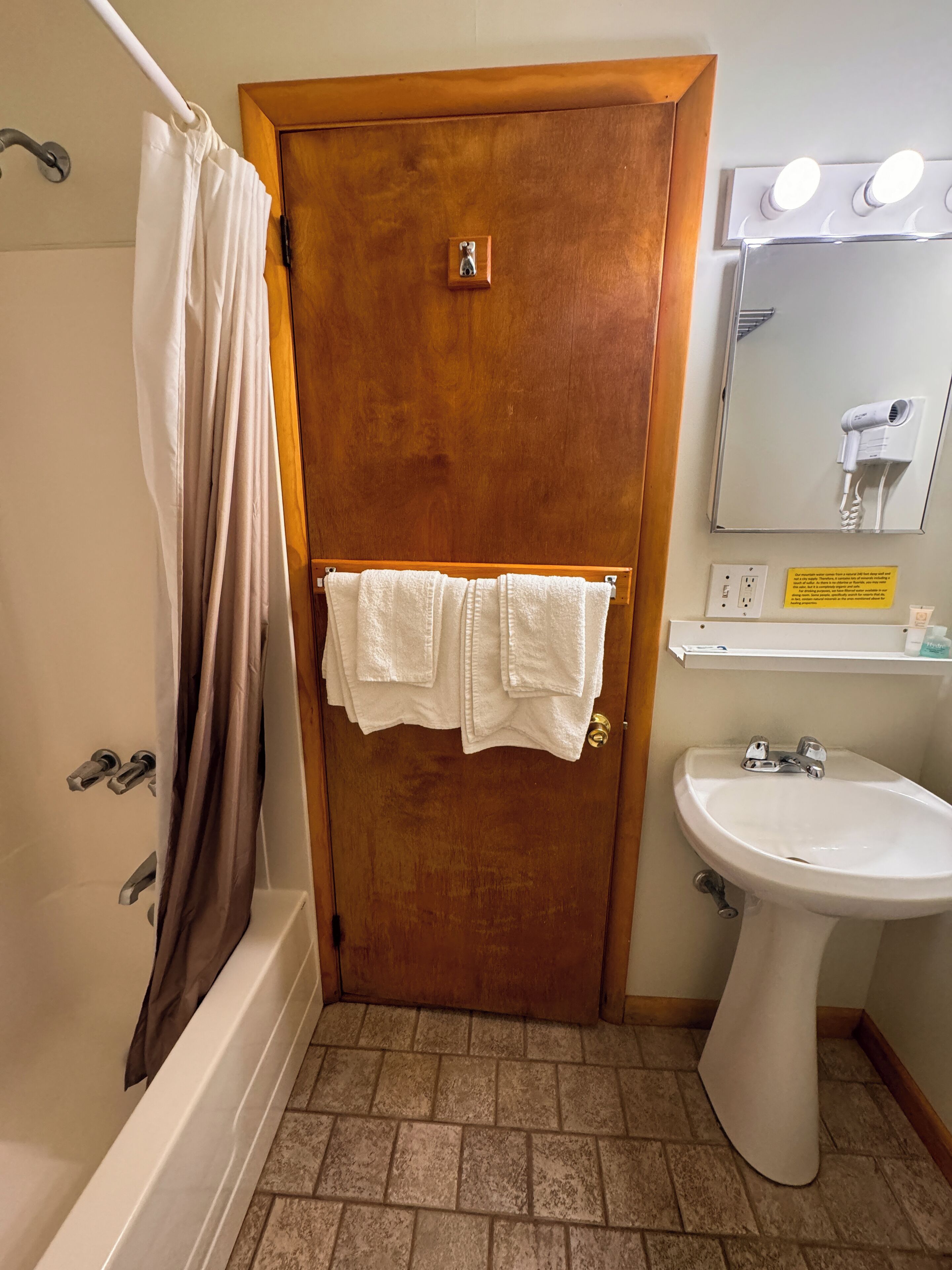 Un baño con puerta de madera, un lavamanos blanco, un espejo con luces y una zona de ducha.