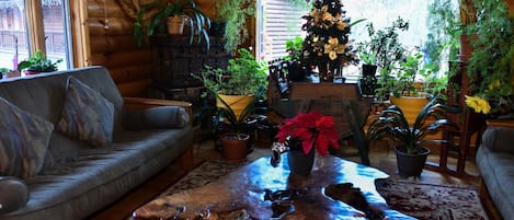 A cozy living room with a wooden log wall, a grey sofa, a coffee table with a unique wooden base, and various potted plants.