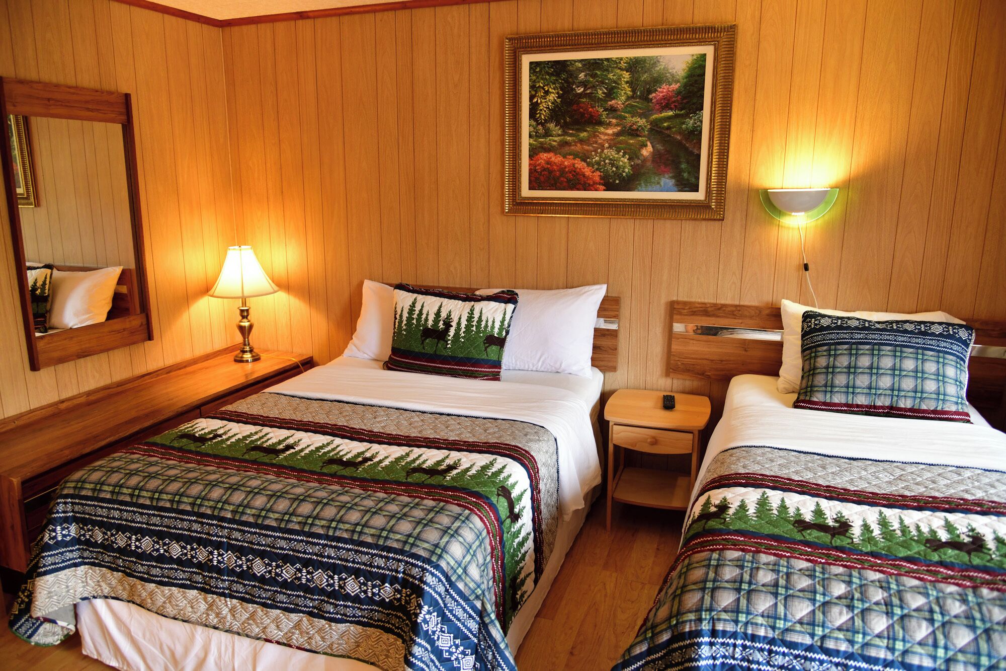 A hotel room with two beds, wooden walls, a painting, and a mirror.