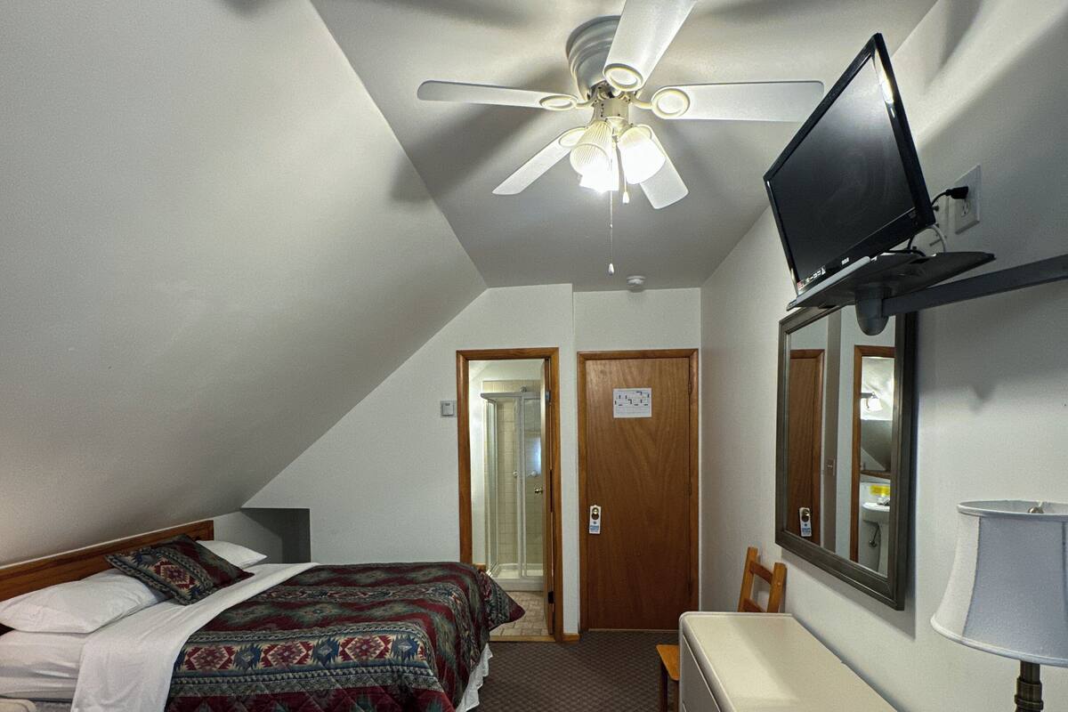Ein Hotelzimmer mit zwei Betten, einem Deckenventilator, einem an der Wand montierten Fernseher, einem Schreibtisch mit Lampe und einem Spiegel.