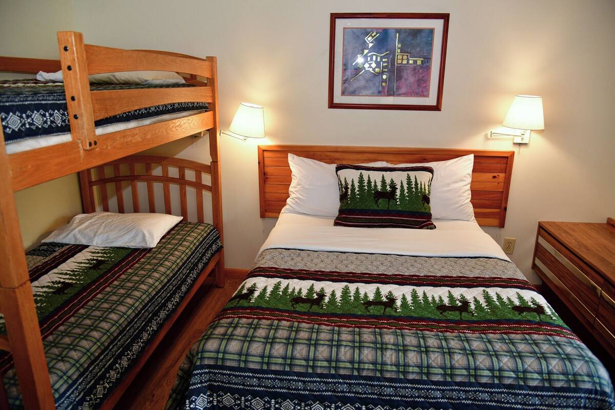 Ein Zimmer mit Etagenbett, zwei Einzelbetten, einem Kopfteil mit einem Gemälde und zwei Nachttischlampen.