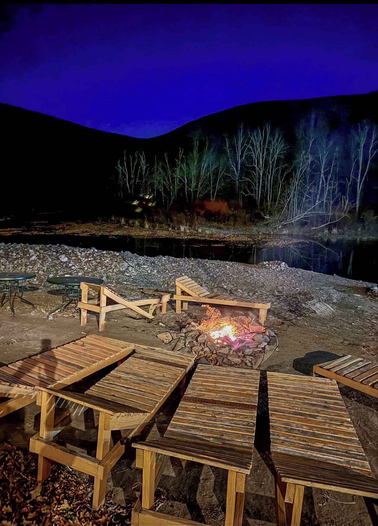 A campfire surrounded by wooden benches and tables, set against a backdrop of a river and trees.