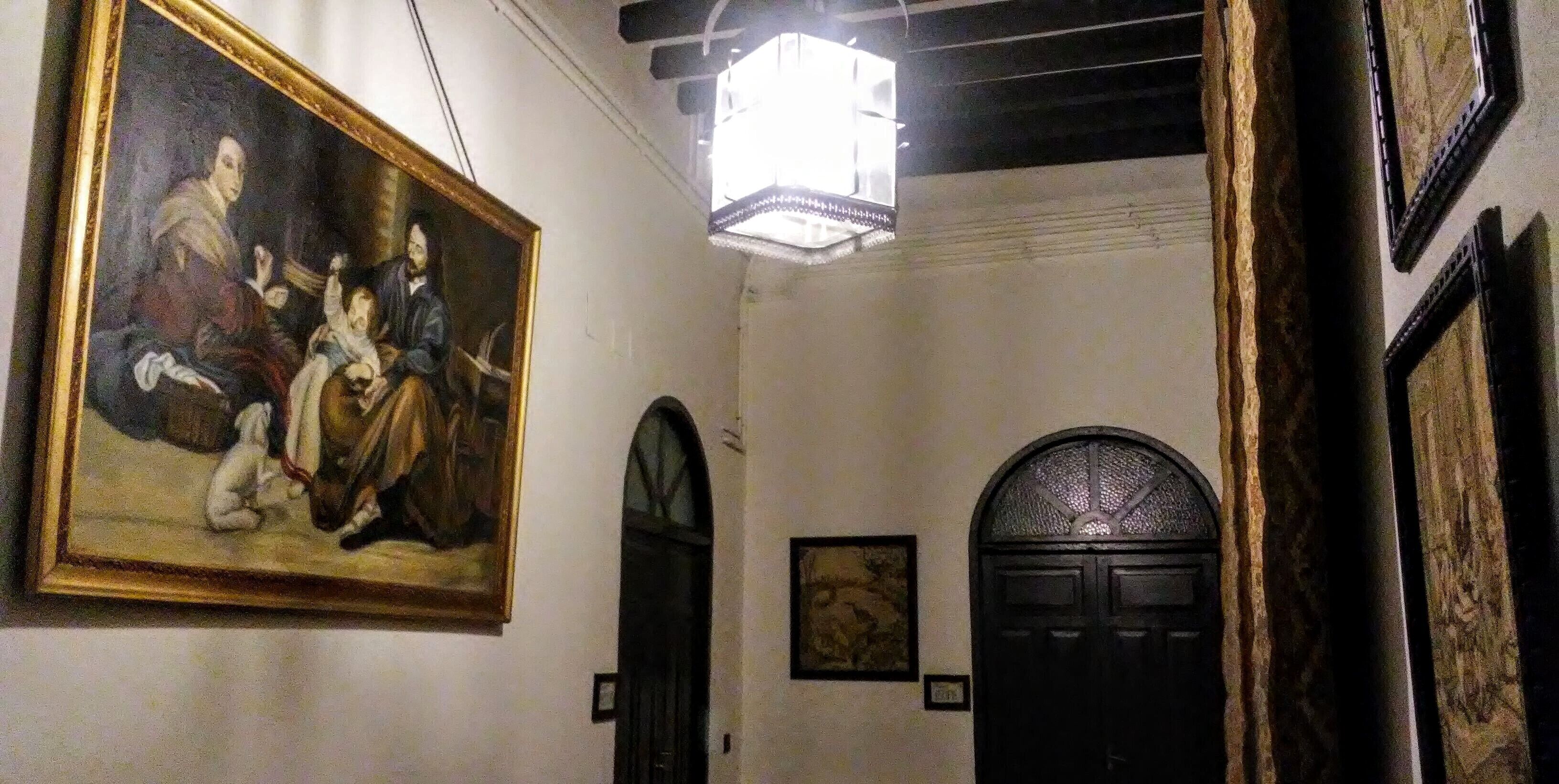 A hallway with a classical painting, a dark arched door, and a framed picture on the wall.