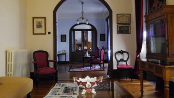 A traditional living room with a wooden floor, a glass coffee table, and red upholstered chairs. There is a radiator on the left and a wooden cabinet on the right.