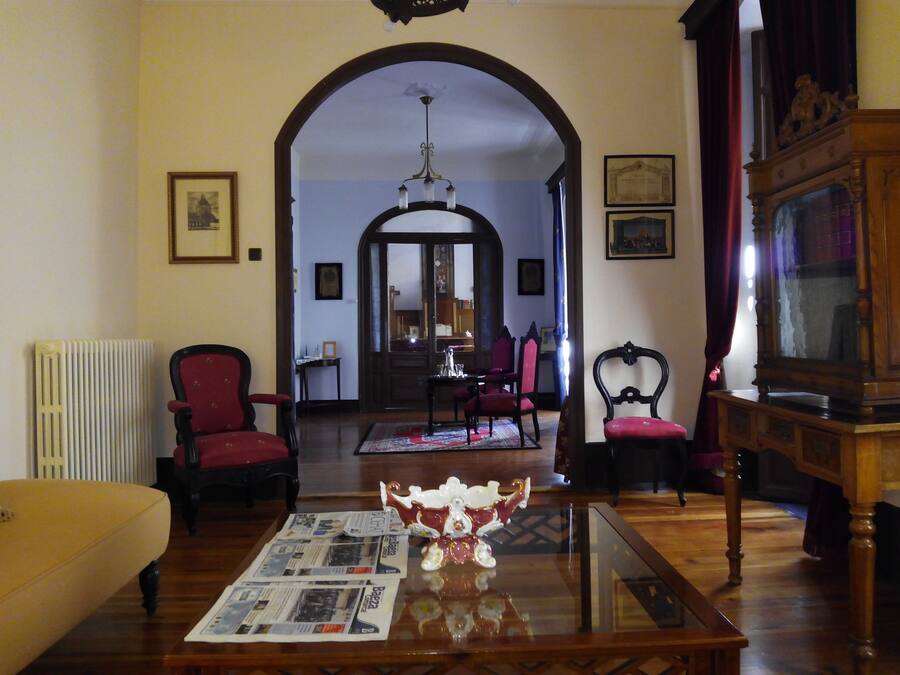 A traditional living room with a wooden floor, a glass coffee table, and red upholstered chairs. There is a radiator on the left and a wooden cabinet on the right.
